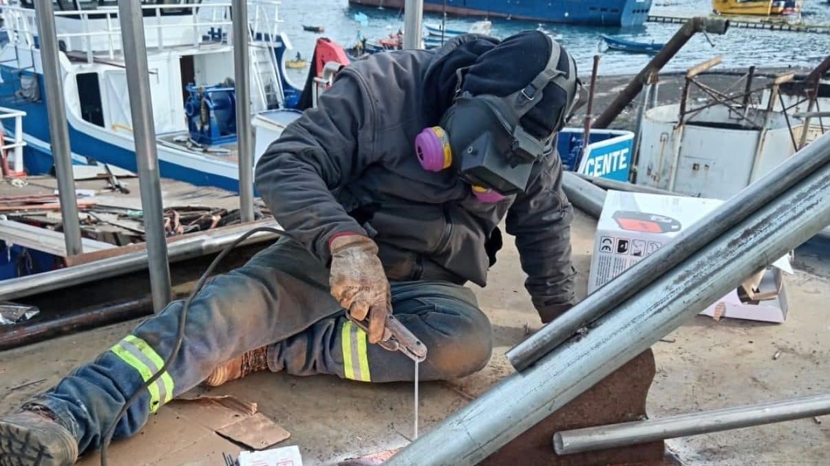Soldador haciendo mantención de estructura sobre el muelle, con la flota pesquera de Talcahuano detrás